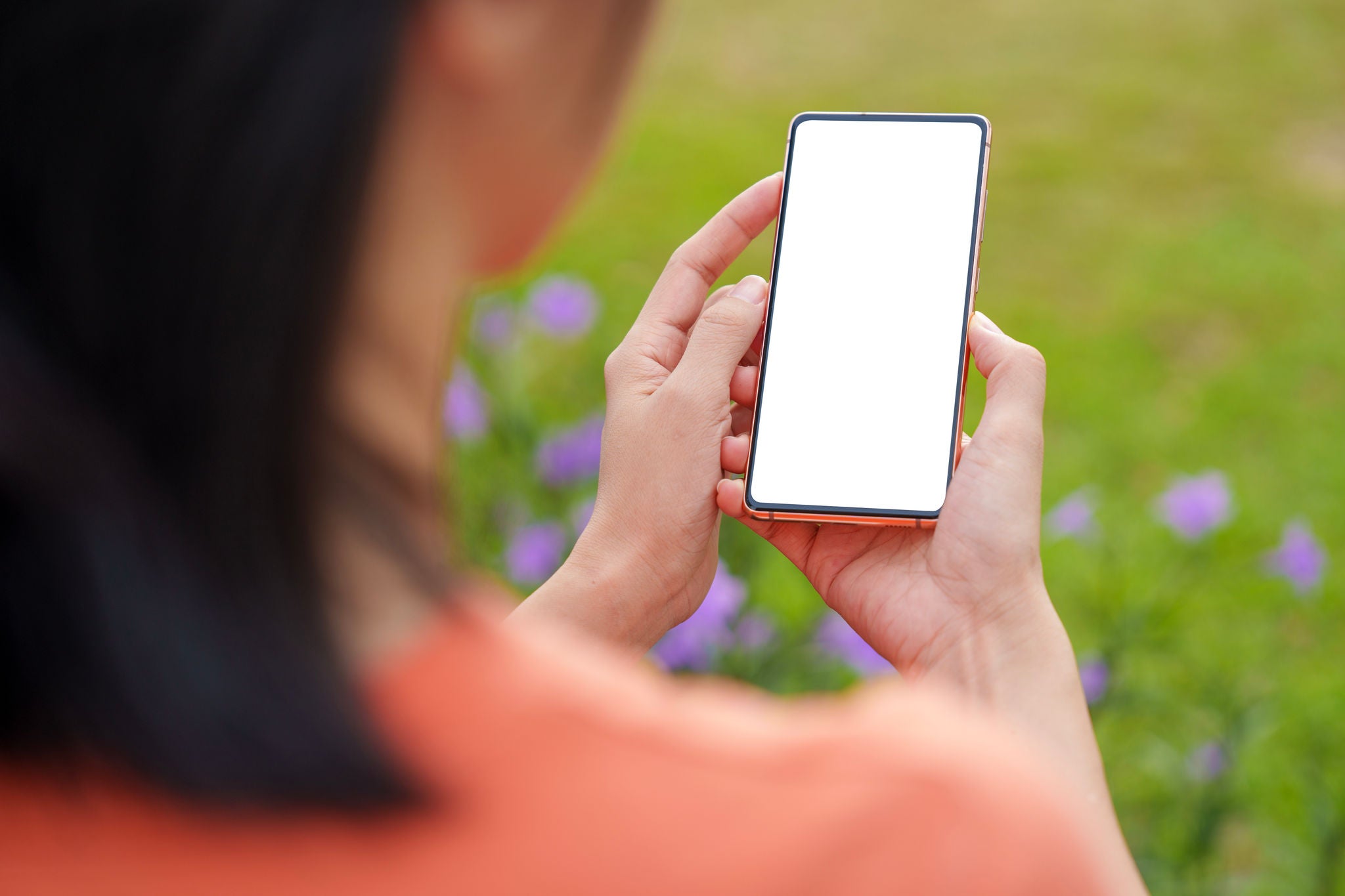 Over the shoulder view of an Asian woman using a smartphone with a blank screen while sitting in a public park.