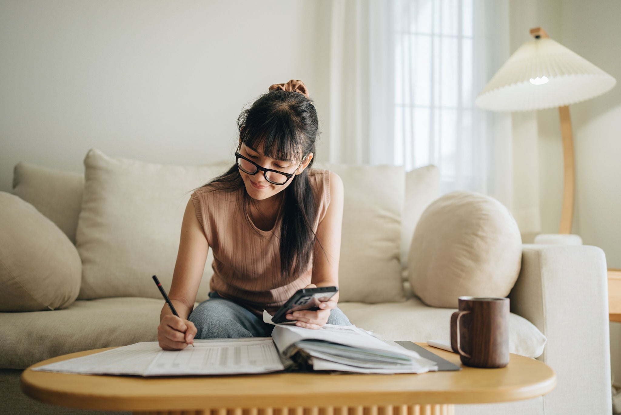 A woman sits at her living room with smartphone and financial reports doing her monthly budget.