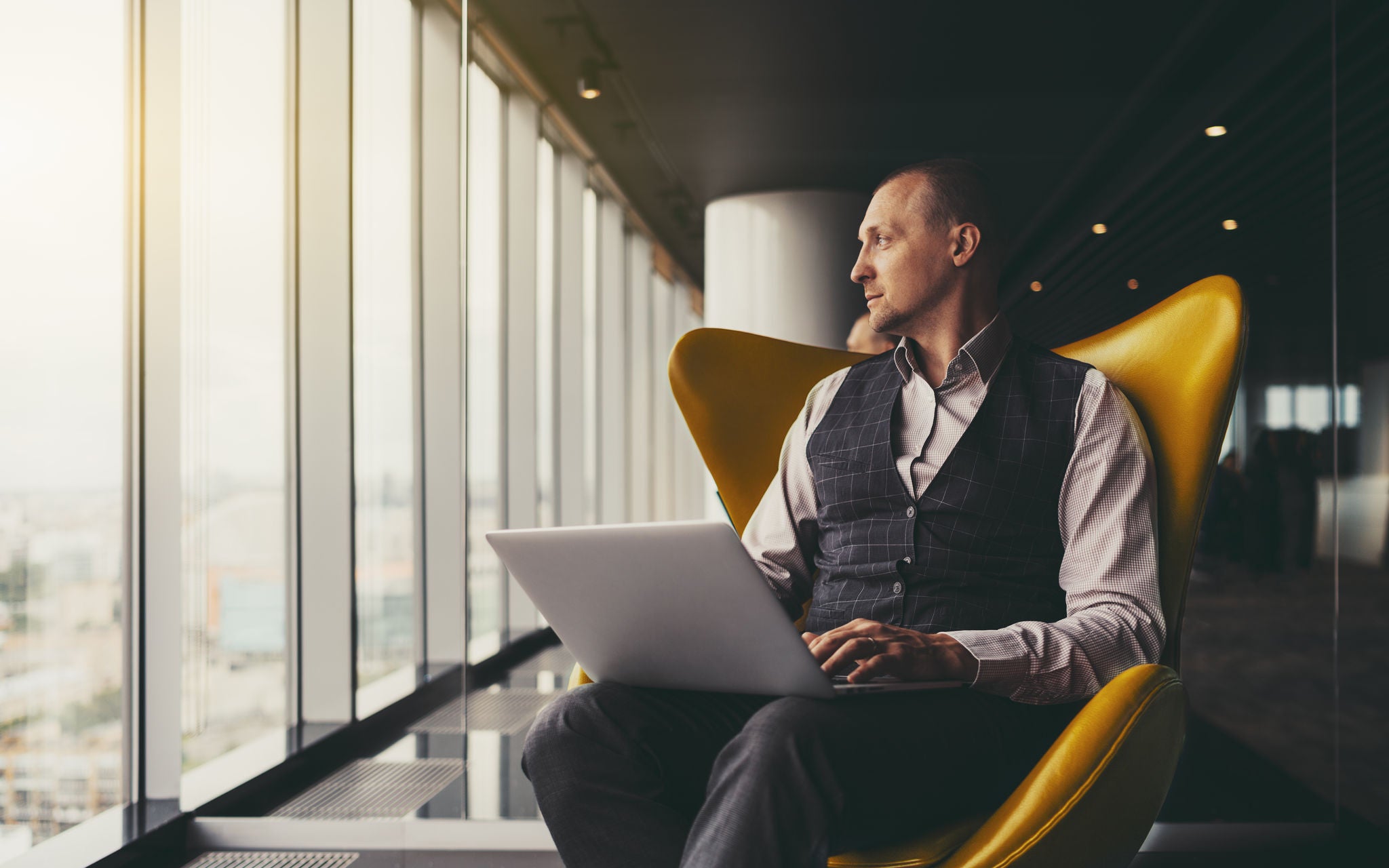 A confident relaxed successful mature man entrepreneur sitting with a laptop on an orange armchair and thoughtfully looking aside on an urban skyline outside the window; a copy space place on the left