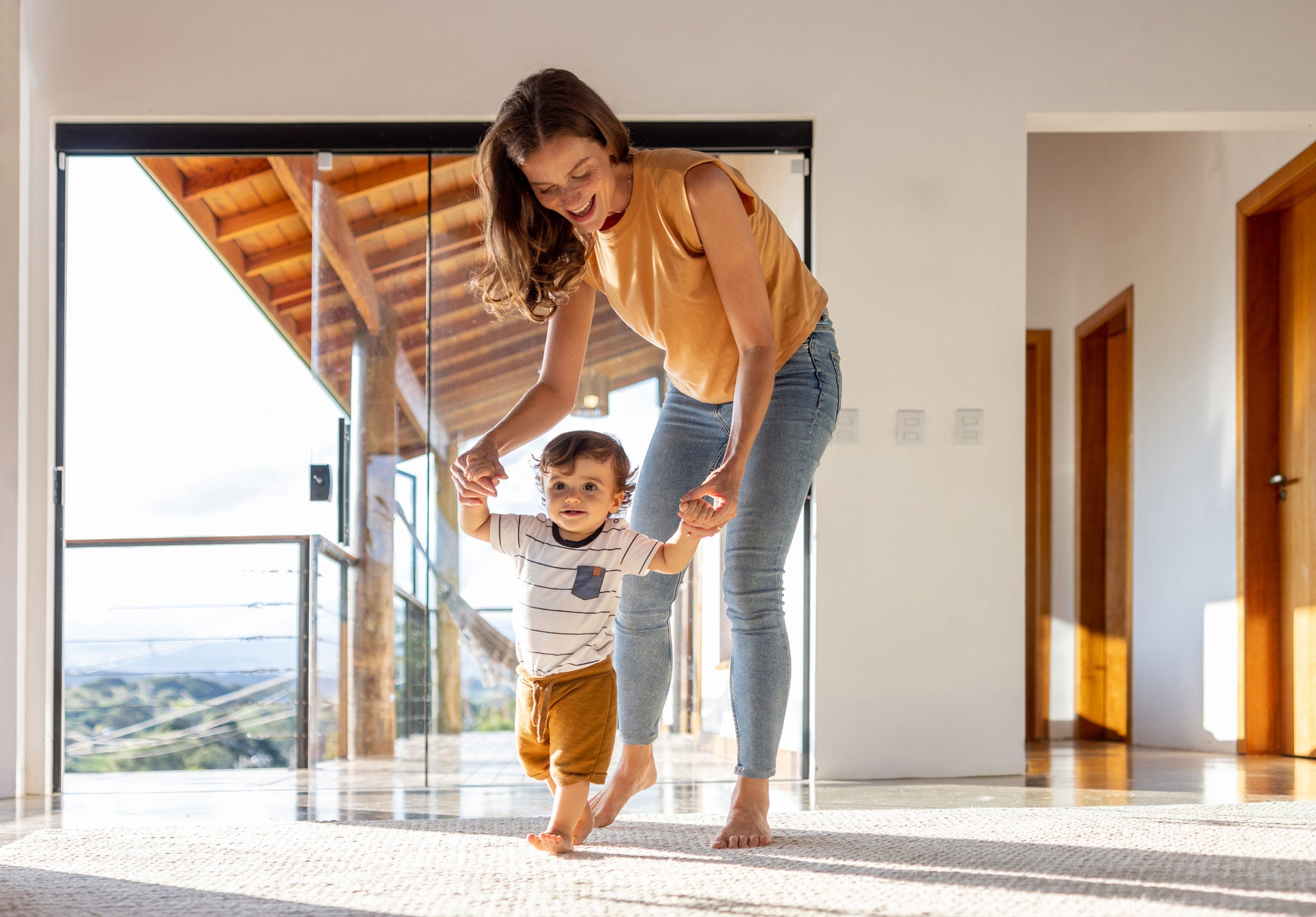 Happy toddler at home learning how to walk with the help of his mother - first steps concepts